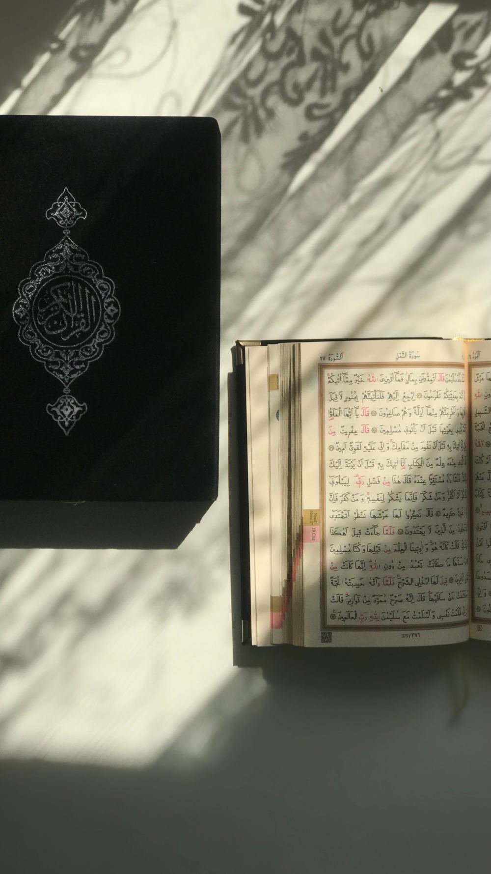 A mushaf open beside its cover, leaf shadows falling across the page