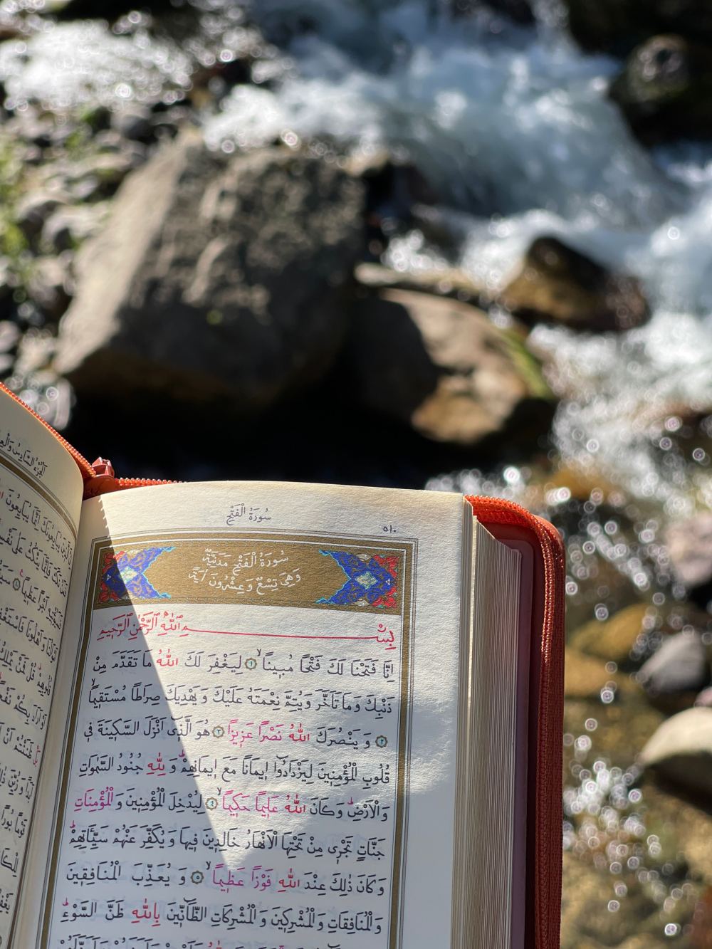 A mushaf open at Surah al-Fath beside a mountain stream
