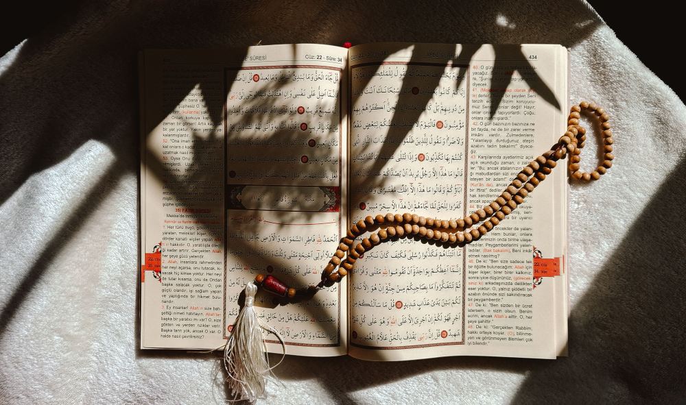 An open mushaf with a string of prayer beads across it, in dappled light